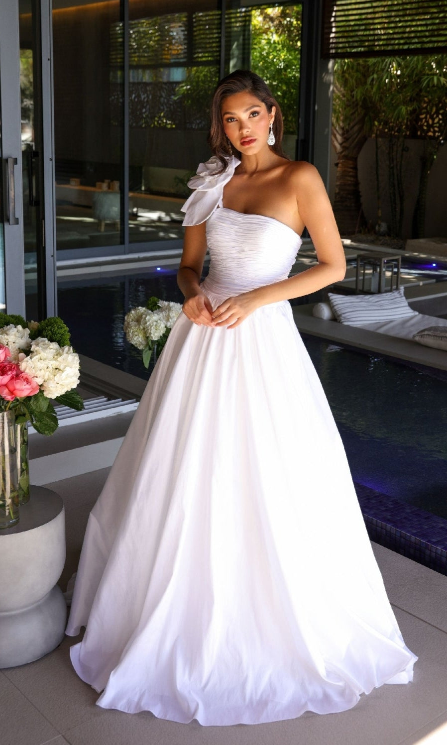 A woman stands indoors by a pool, wearing the Ava Presley 26524 long prom dress—an elegant white, one-shoulder gown with a large floral detail. A flower arrangement sits on a small round table nearby.